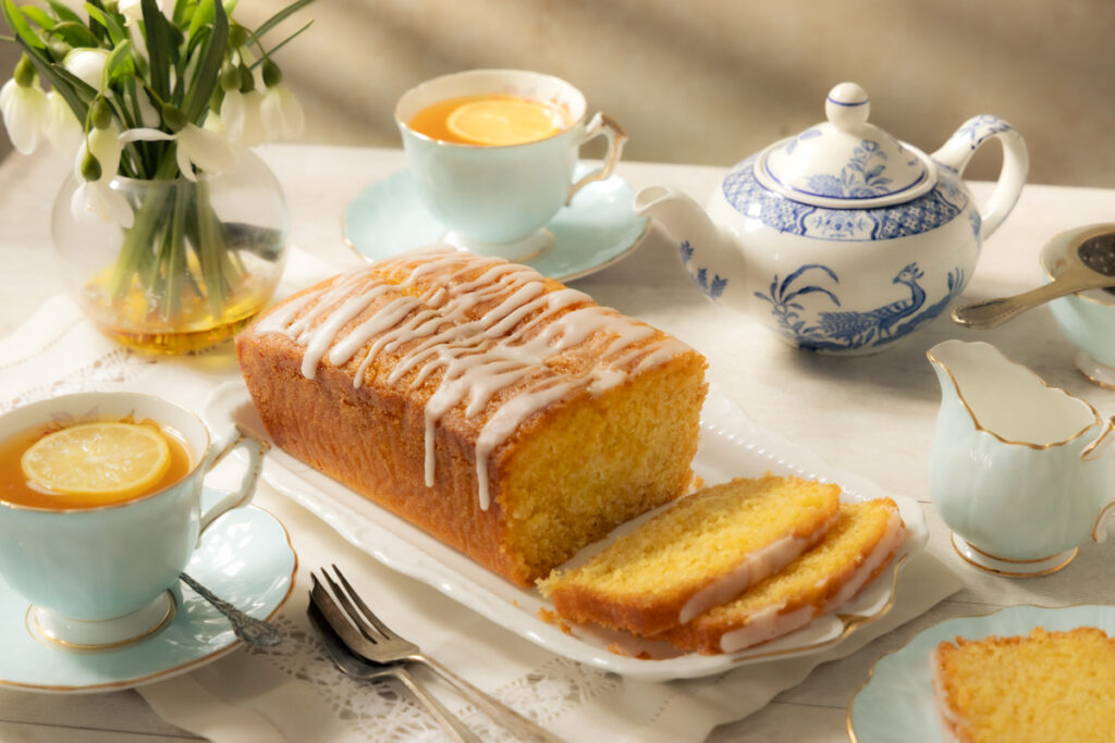 Lemon drizzle loaf cake on a pretty vintage serving plate on a table set for afternoon tea with vintage teapot and cups and saucers with tea and lemon slices